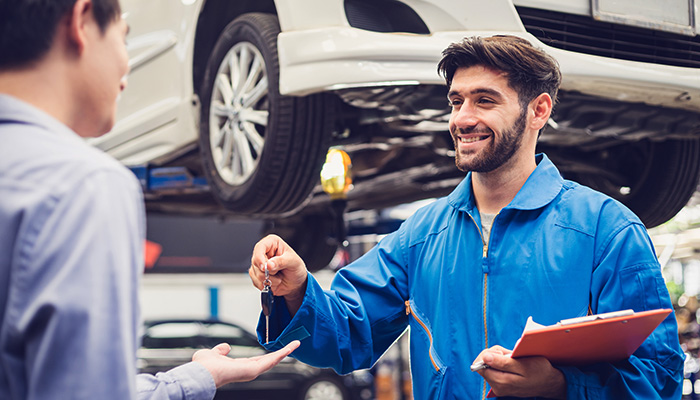 GMC Certified Service technician in front of a white SUV up in the air behind him giving keys to the owner in the shop after servicing his SUV