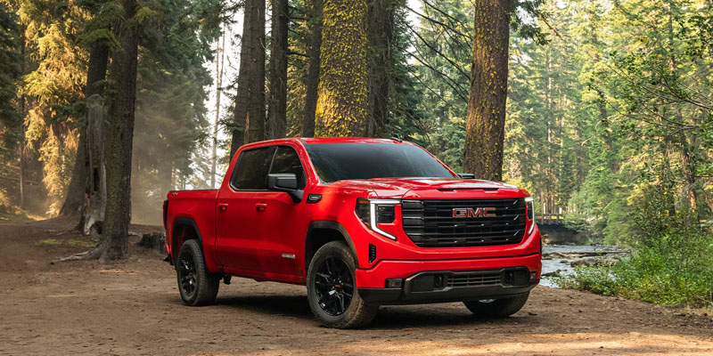 A red 2026 GMC Sierra 1500 truck is parked beside a wide creek in a forest park as the sun rays light it up.